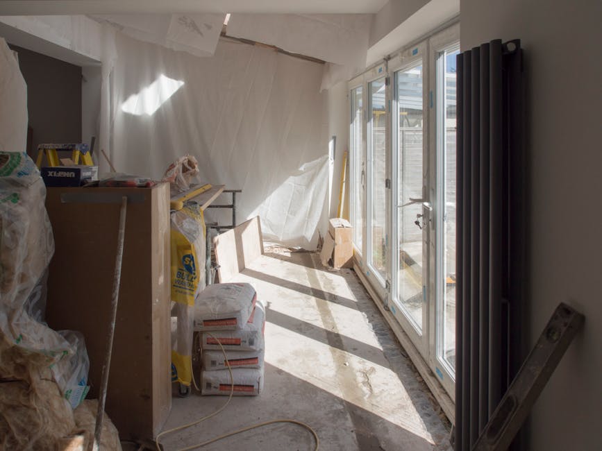 The image shows an interior space undergoing renovation, with construction materials and tools arranged on the floor and surfaces. Bright natural light enters through large glass sliding doors, which are partially open, revealing an outdoor area. The room has a concrete floor that appears to be in the process of cleaning or floor preparation. A black radiator is mounted on the wall to the right, and a white curtain or sheet hangs loosely from the ceiling to the left. Various construction supplies, including bags of cement or plaster, boxes, and small tools, are visible on and around a wooden work surface. The space appears to be in the midst of a deep cleaning process following renovation work, aligning with the services provided by Cleaners Highbury, focusing on post-build sanitisation, surface cleaning, and dust removal to prepare the area for final finishing.