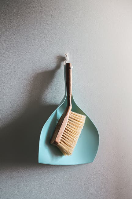 A cleaning tool set hanging on a white textured wall, consisting of a light blue dustpan and a wooden-handled scrub brush with beige bristles. The dustpan is positioned vertically with the handle pointing downward, while the scrub brush is resting inside the dustpan with its handle angled slightly to the left. The scene is illuminated with soft natural light, highlighting the clean and tidy appearance of the surfaces, suitable for surface cleaning, deep cleaning, or sanitisation as part of domestic cleaning services offered by Cleaners Highbury.