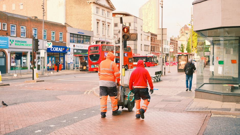 Two street cleaners wearing high-visibility orange uniforms are pushing a cleaning cart on a city sidewalk in Highbury, near a bus stop with a red double-decker bus in the background. The area is well-lit with natural daylight, and the surroundings include various shops, such as a Yorkshire Building Society branch and a store named Coral, along with pedestrians walking past. The pavement is clean, and the scene captures an urban setting focused on maintaining hygiene and cleanliness, representative of the professional cleaning services offered by Cleaners Highbury, as referenced on the page about avoiding hidden cleaning charges in Highbury and N5.