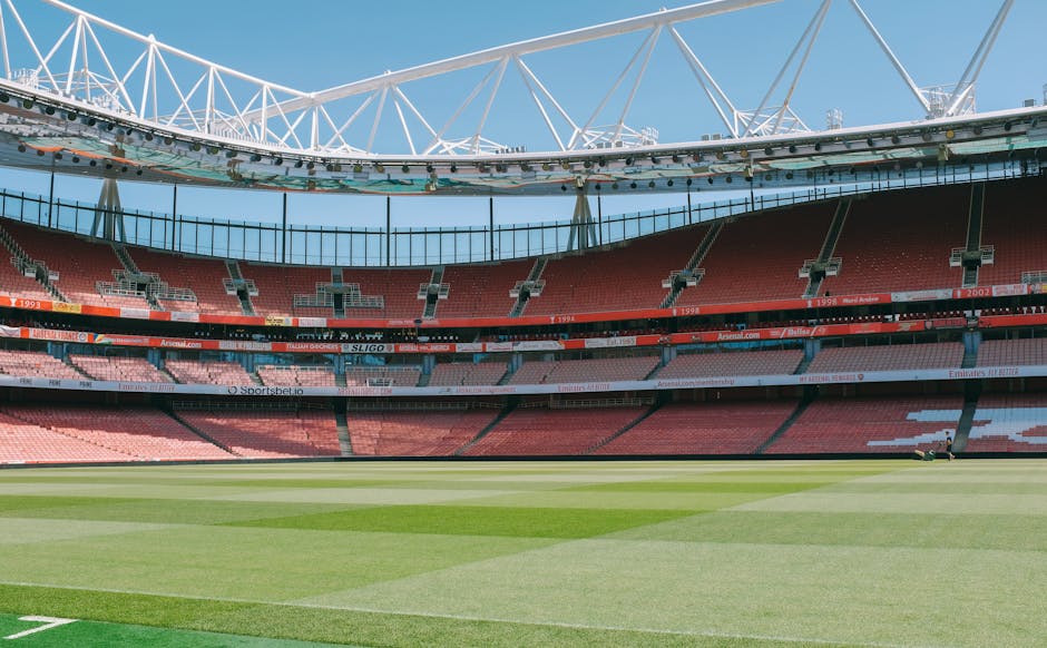 Inside a large, modern stadium with a well-maintained grass pitch, showing multiple tiers of red and pink seating areas, clean and orderly. The upper structure includes a white, intricate roof framework that extends over the seating sections, with clear blue sky visible through the open sections. The playing surface is lush, even, and free of debris, indicating professional upkeep. The stadium lighting fixtures are attached to the roof structure, and the overall environment appears bright and pristine, reflecting high standards of cleanliness and maintenance suitable for sports events. Mentioned here is the importance of surface cleaning and sanitisation that Cleaners Highbury provides for commercial spaces like stadiums, aligned with the high-quality standards of deep cleaning and hygiene required in such large public venues.
