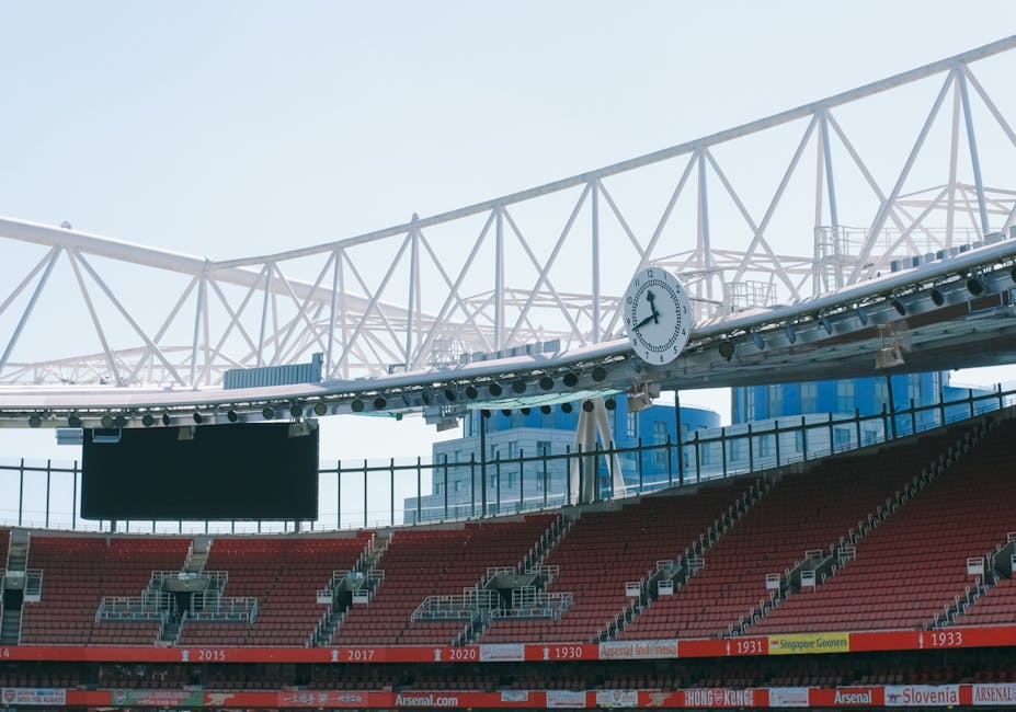 An empty football stadium with a well-maintained grass pitch featuring white boundary lines, surrounded by rows of orange and grey seating sections under a large, white arched roof structure supported