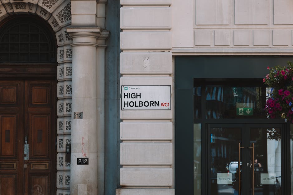 Close-up photograph of the exterior wall of a building on High Holborn, showcasing a white sign with black text reading 'HIGH HOLBORN WC1' situated between a decorative stone column and a large glass door. The building features a beige stone facade, with the door framed by a dark green panel. To the right of the sign, a metal flower basket filled with vibrant pink and purple flowers hangs beside the glass entrance. The glass door has a green emergency exit sign above it, and the reflection in the glass shows a glimpse of parked cars and street activity. The overall scene emphasizes a clean, well-maintained urban environment with professional cleaning services provided by Cleaners Highbury, as referenced on the company’s website for office and retail cleaning in Highbury.