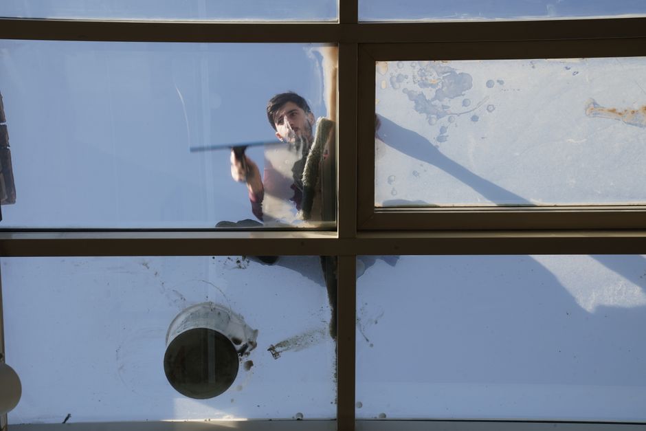 A window cleaner is seen through a large glass window, holding a squeegee in their hand and in the process of cleaning the glass surface. The window features a metal frame dividing it into multiple se